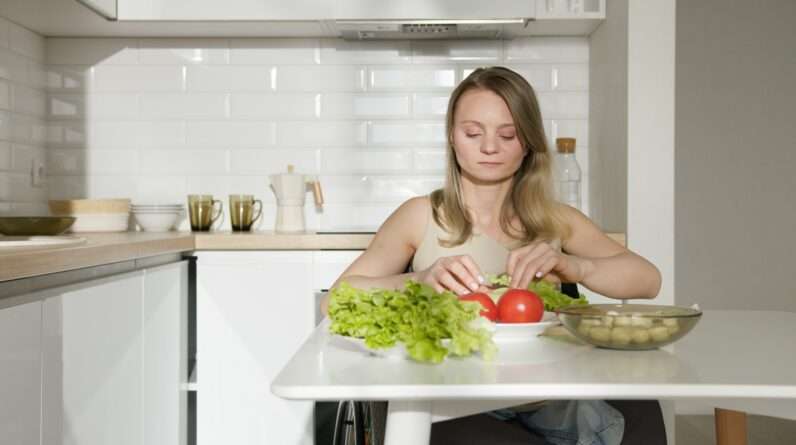 A Woman in Beige Tank Top Sitting on the Kitchen Near the Table With Vegetables