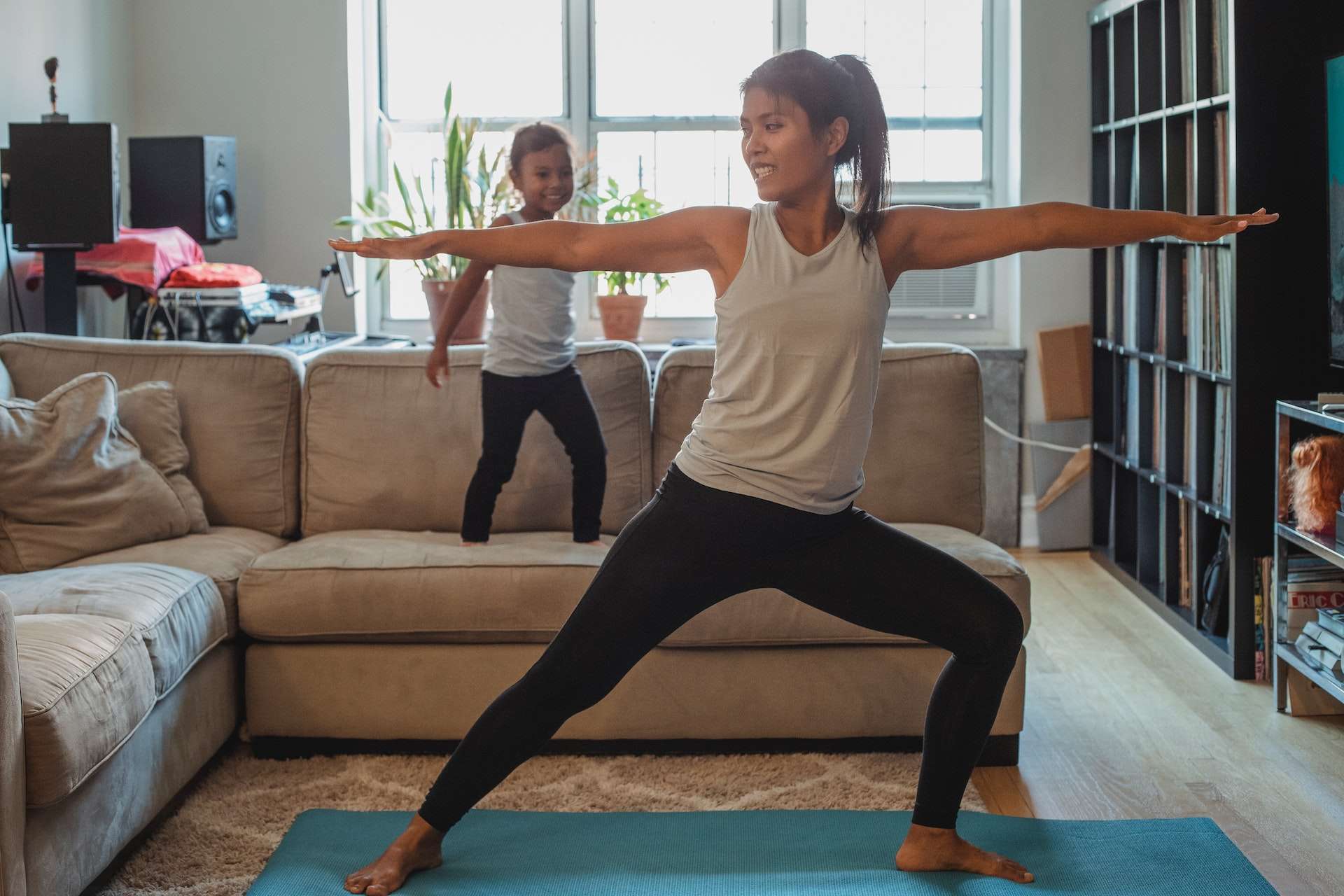 Ethnic woman performing Warrior Pose at home near adorable daughter 