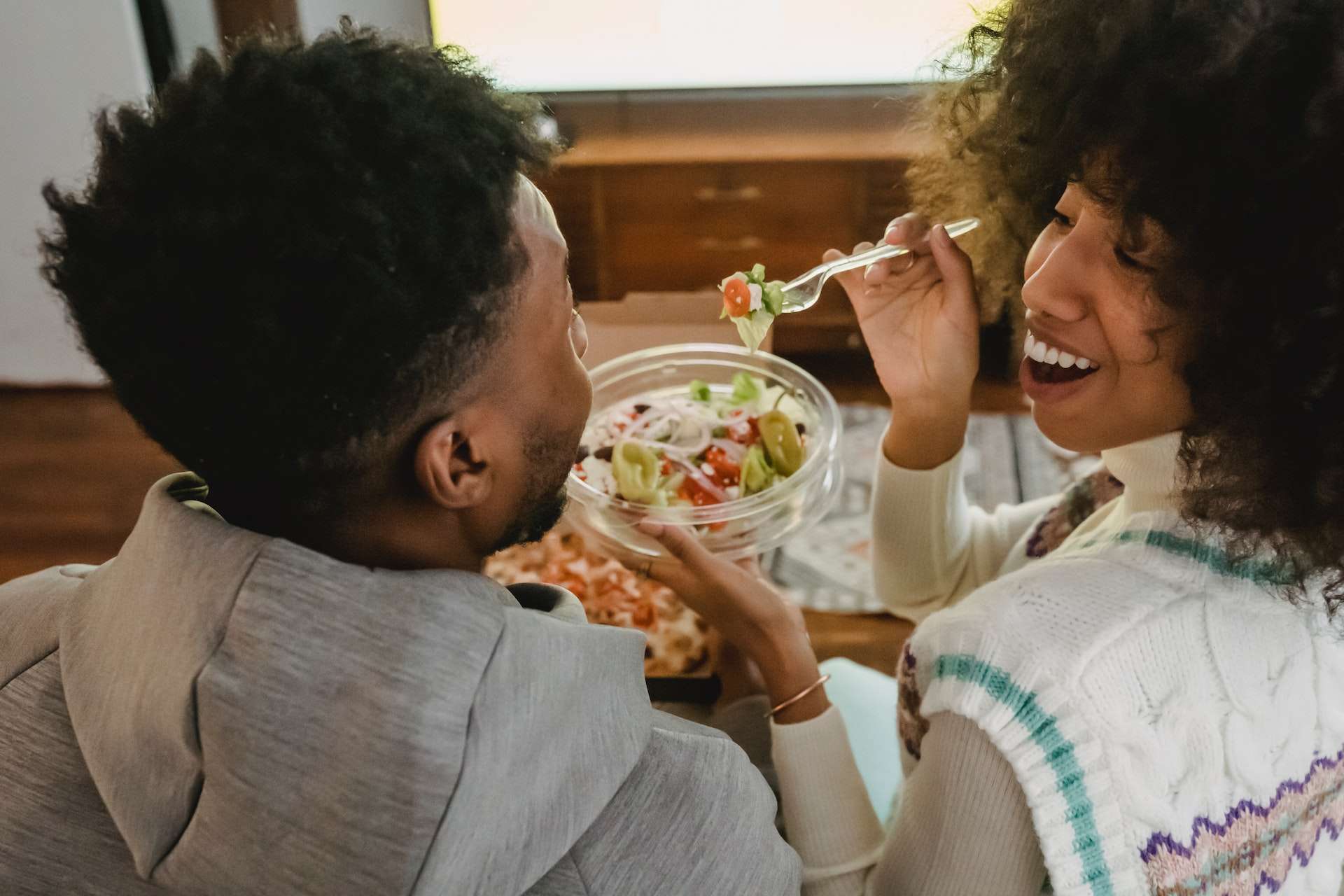 Happy young ethnic woman feeding boyfriend with salad while sitting on couch 