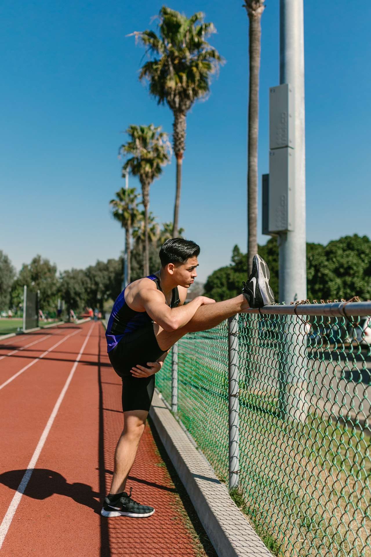 Man in Black Sportswear Stretching Leg Beside Wire Fence on Track Field 
