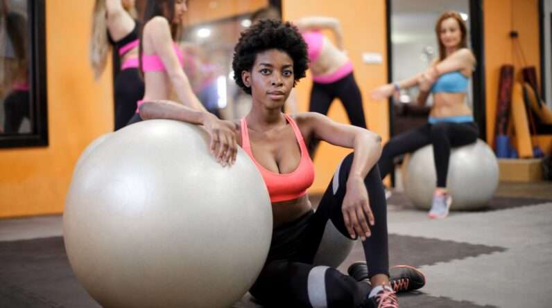 Young ethnic woman with fit ball sitting on floor while training in modern gym