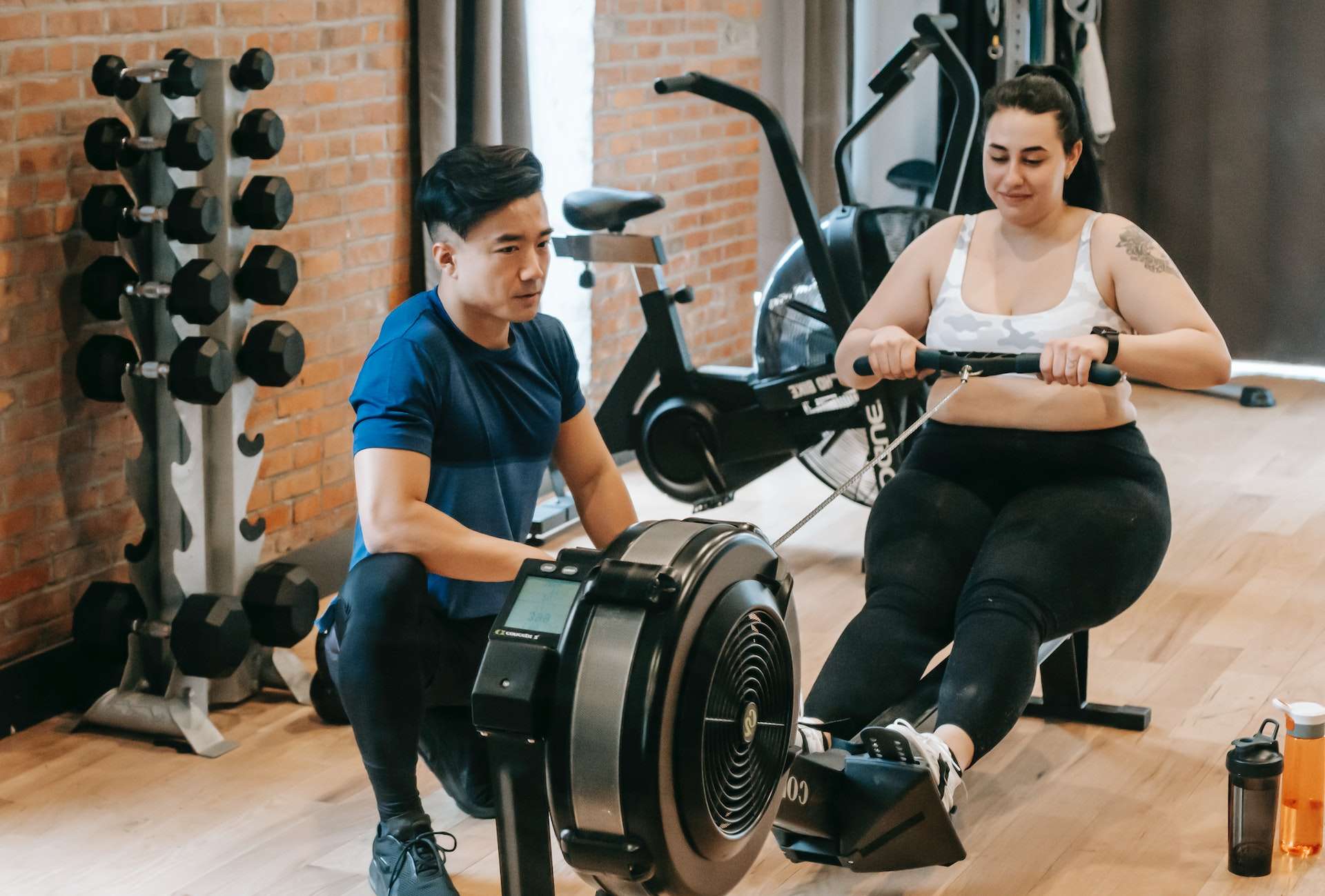 Happy young overweight woman exercising on rowing apparatus during fitness training with Asian instructor
